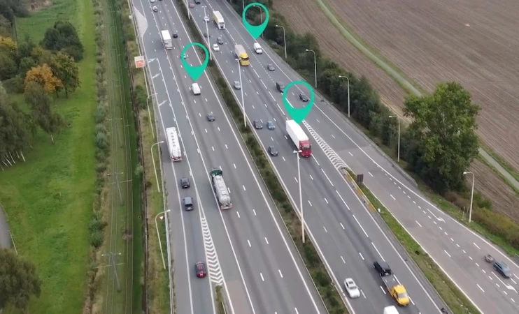 A busy motorway with cars, trucks, and vans driving on it with green location pinpoint icons above three vehicles.